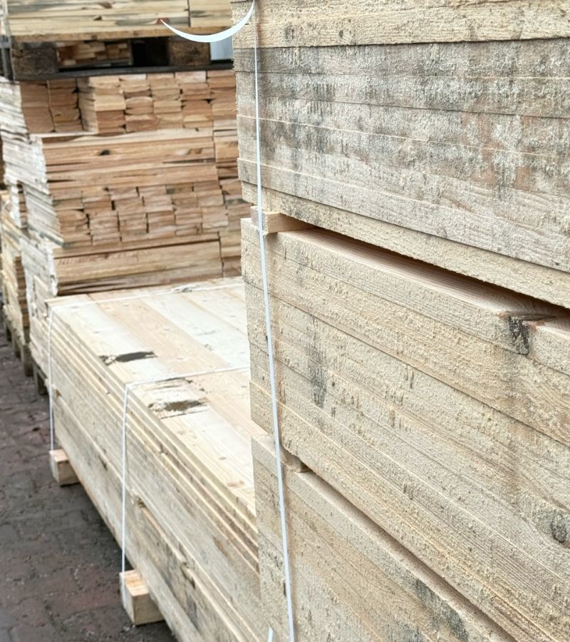 Zbliżenie na stosy surowych desek drewnianych przeznaczonych do produkcji palet, spiętych białą taśmą, składowanych na zewnętrznym placu magazynowym.