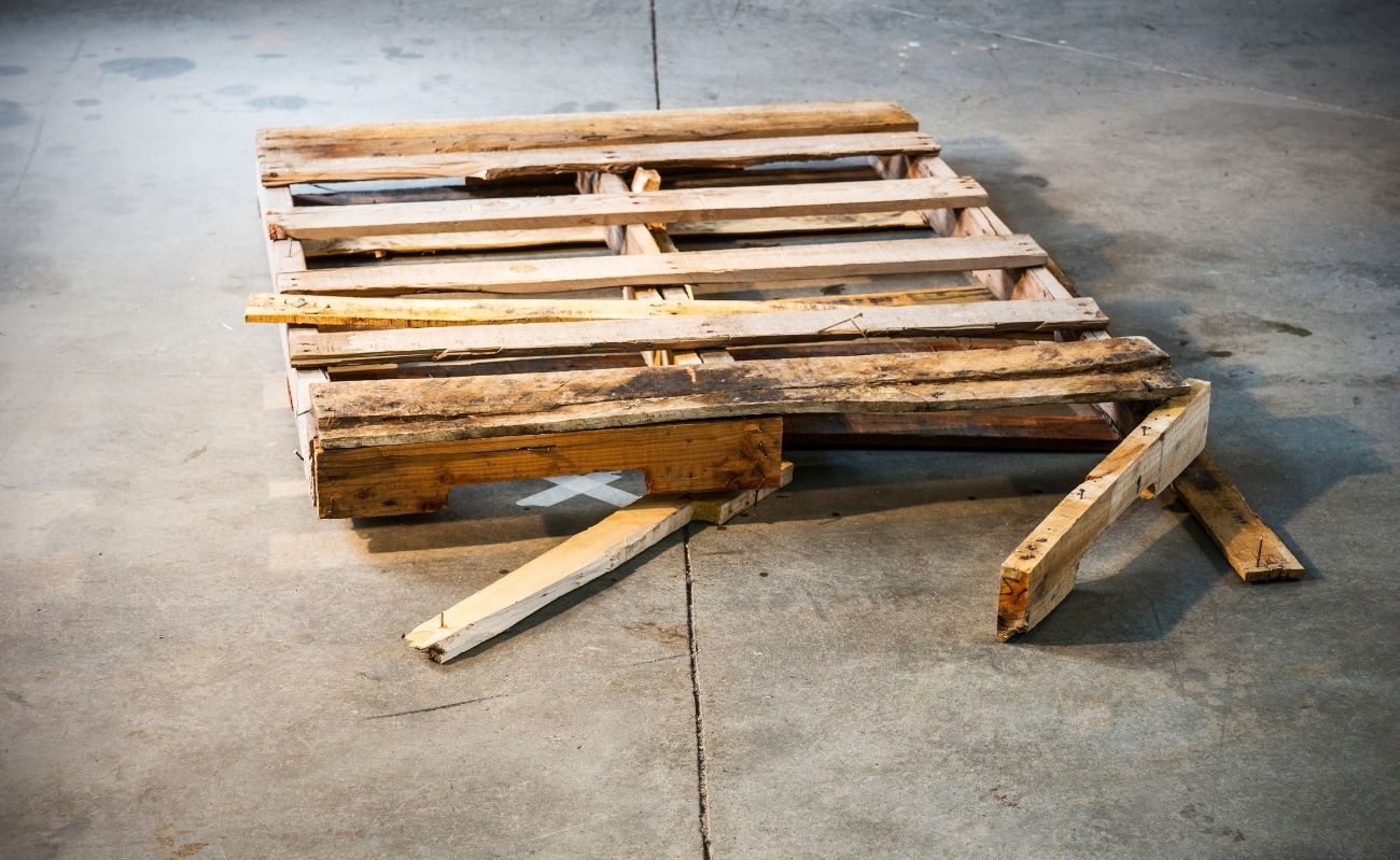 Stara, uszkodzona paleta drewniana z połamanymi deskami na betonowej podłodze, przeznaczona do profesjonalnej renowacji lub recyklingu