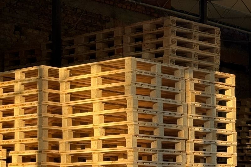 Zbliżenie na stos nowych, jasnych palet drewnianych w magazynie, z widocznymi w tle wysokimi stosami używanych palet z oznaczeniami EPAL.