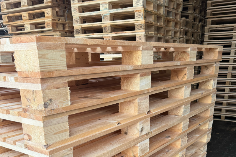 Zbliżenie na stos nowych, jasnych palet drewnianych w magazynie, z widocznymi w tle wysokimi stosami używanych palet z oznaczeniami EPAL.