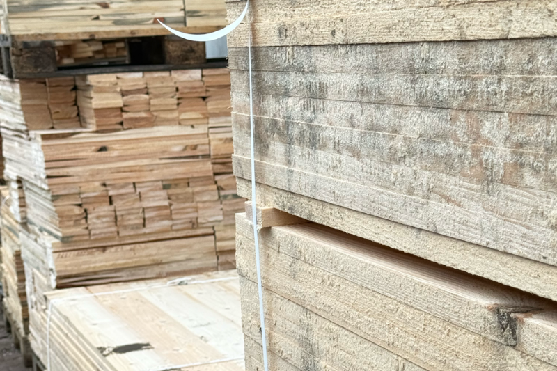 Zbliżenie na stosy surowych desek drewnianych przeznaczonych do produkcji palet, spiętych białą taśmą, składowanych na zewnętrznym placu magazynowym.