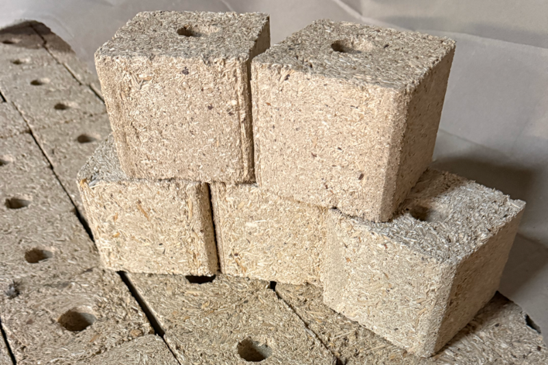 Zbliżenie na kwadratowe wsporniki (klocki) do palet wykonane z prasowanych wiórów drzewnych z widocznym otworem montażowym, ułożone jeden na drugim.