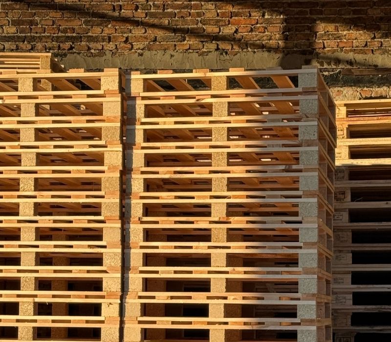 Stosy nowych, jasnych palet drewnianych ułożone przy ceglanej ścianie magazynu w świetle zachodzącego słońca.
